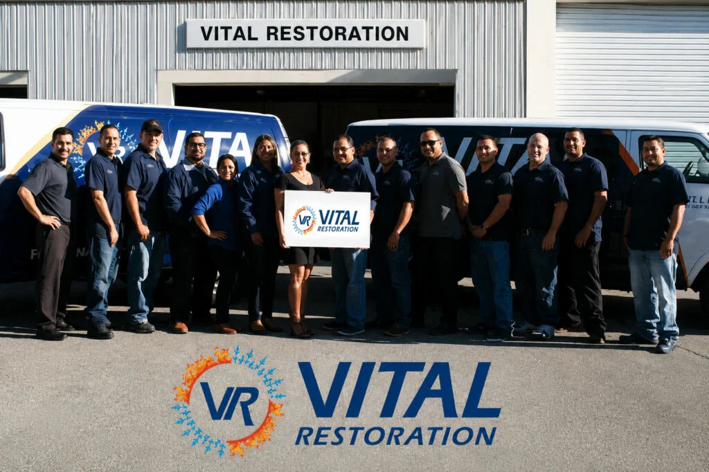 A group of people in dark uniforms stand in front of two Vital Restoration disaster restoration franchise vans and a building.